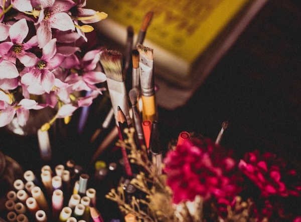 Creativity, art. Close up of paint brushes and pencils in a holder on a desk with flowers around.