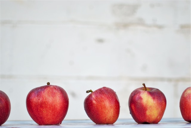 Red apples lined up against a white wall. Different shapes, some tilted, not perfect. Perfection is not necessary to progress steadily.