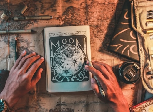 Book with artistic full moon illustration at the center of a table. Hands holding it, with pen in one hand as if continuing to write and illustrate. Set intentions with new moon energy.