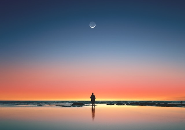 Person standing on a beach facing the water, twilight sky, and a sliver of a new moon.