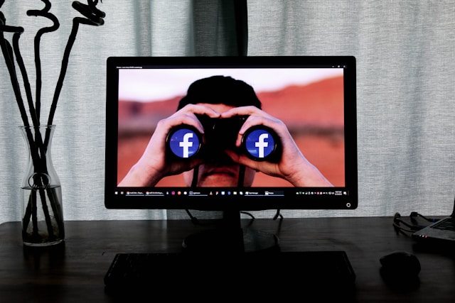 Computer monitor on a desk. On the screen is the headshot of a man facing us, looking through binoculars with facebook logo on it. Prying, privacy breaching energy. 