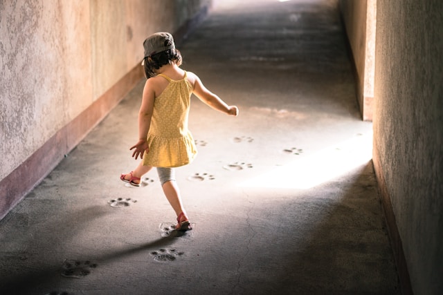 little girl in yellow dress and dark cap playing by herself. Lost in her game. Back turned to camera. Large paw prints on the floor, she's skipping from one print to the next.