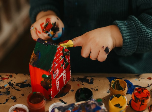 Close up of little kid hands finger painting a red wooden toy house. Lost in creative flow state.