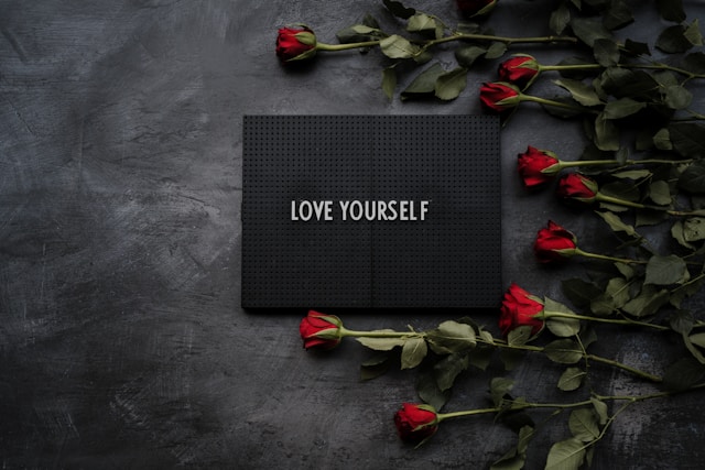 Dark background with red, long stemmed roses layed on one side. Black board with words "love yourself" printed on it.