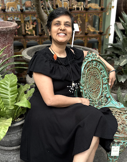 Widely smiling Indian lady with short hair, Susmitha (vegan business coach), in a black midi dress. She's sitting on a short verdigris metal garden chair, arm draped on the back of another chair next to her. Looking directly at the camera.