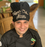 Indian man in black checkcoat and chef hat. Looking at the camera and smiling. Vegan Chef Ram.