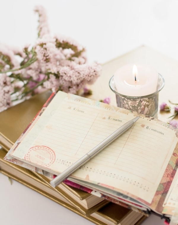 Daily planner open with soft pink flowers and candle around it. Pen on the planner. Photo has a gentle, easy energy. The magic of scheduling your life in alignment with your nature, according to who you are.