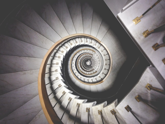 Photo of spiral while stairs with golden brown bannister captured from above. Representing the emotional spiral of going up or down.