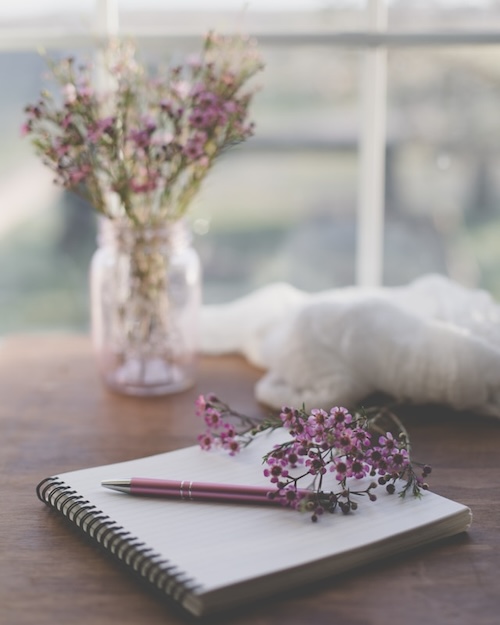 Open book with pen and flowers on it. Glass jar with flowers near it on the wooden desk. Journaling is a high quality leisure activity.