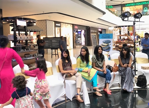 Four Indian ladies with long hair sitting on chairs covered with white sheets in the middle of a mall.