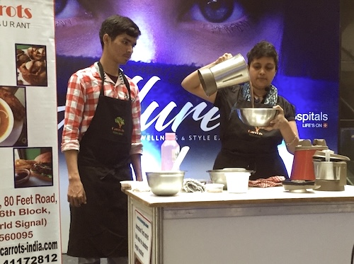 Short haired Indian lady (Susmitha Veganosaurus) pouring vegan chocolate mylk from blender jar to a steel bowl. Next to her a young Indian boy (Jay Thapa) standing in wait to assist her. A collaboration event that flopped because of a lack of crowd turn out.