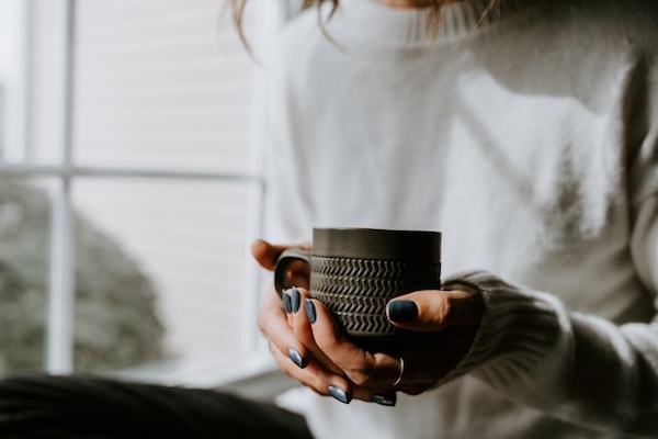 Woman's hands, holding a warm mug. Sunlight and shadows from window on the side falling on her white top. Cozy. Chang your life.