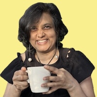 Susmitha Veganosaurus, vegan business coach, chef, multi-passionate creator. Holding a white mug, wearing a black dress, dangly earrings, smiling wide at the camera.