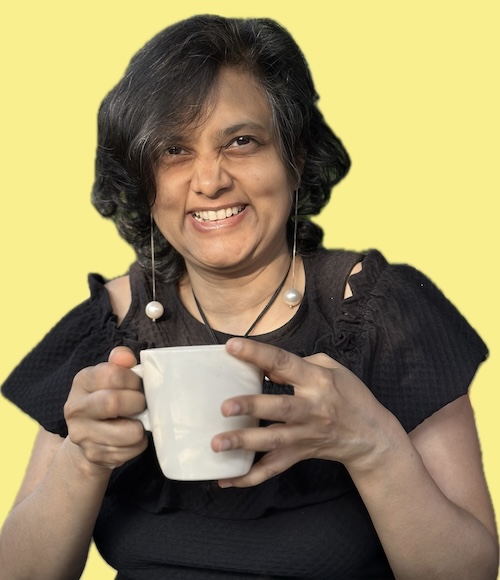 Susmitha Veganosaurus, vegan business coach, chef, multi-passionate creator. Holding a white mug, wearing a black dress, dangly earrings, smiling wide at the camera.