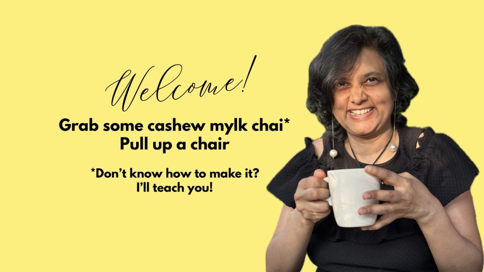 Susmitha Veganosaurus, vegan chef, business coach, spiritual, multi-passionate entrepreneur and teacher. Wearing black dress, dangly earrings, and holding a white mug. Text: Welcome! Grab some cashew mylk chai, pull up a chair. Don't know how to make it? I'll teach you.