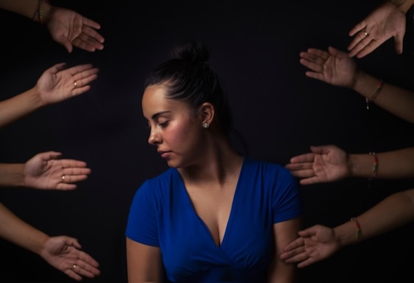 Dark background. Woan in blue dress turning away as uninvited helpful hands cross boundaries and reach out to her in all directions.