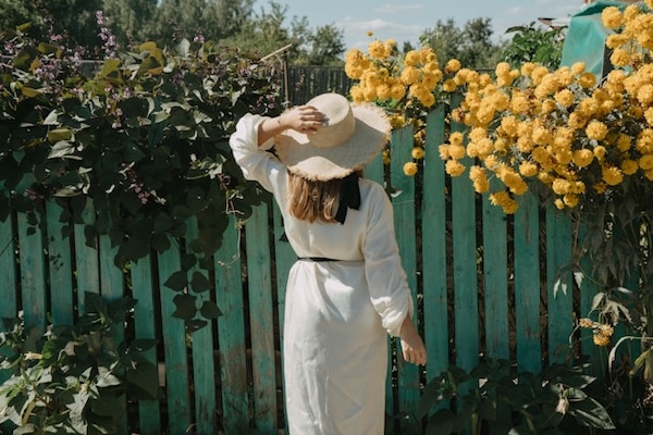 Woman facing away from camera, looking at a wooden fence with flowers and vines on it. She looks to be relaxed, at ease, holding on to her hat.